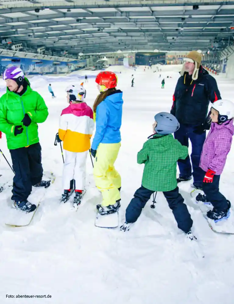 Family in colorful winter gear snowboarding and skiing inside SnowWorld Bispingen – an indoor ski paradise for kids and adults in all weather conditions.