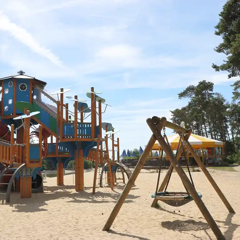 Large adventure playground with slide tower, climbing elements and basket swing on a sandy surface at Südsee-Camp – perfect for families with children, partially accessible.