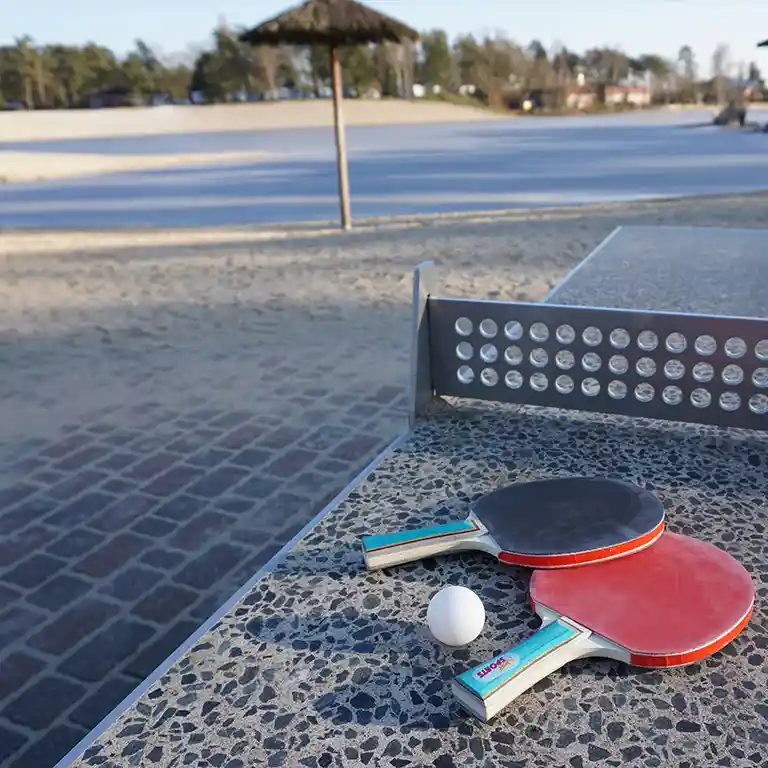 Table tennis paddles and ball on a stone surface in front of a ping pong table at the Südsee-Camp beach with sand and palm-style sunshade in the background