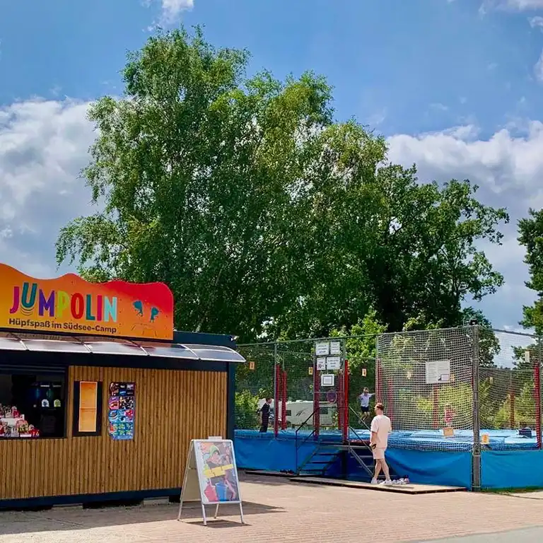 JUMPOLIN trampoline area with kiosk and children jumping at Südsee-Camp, blue sky and trees in the background