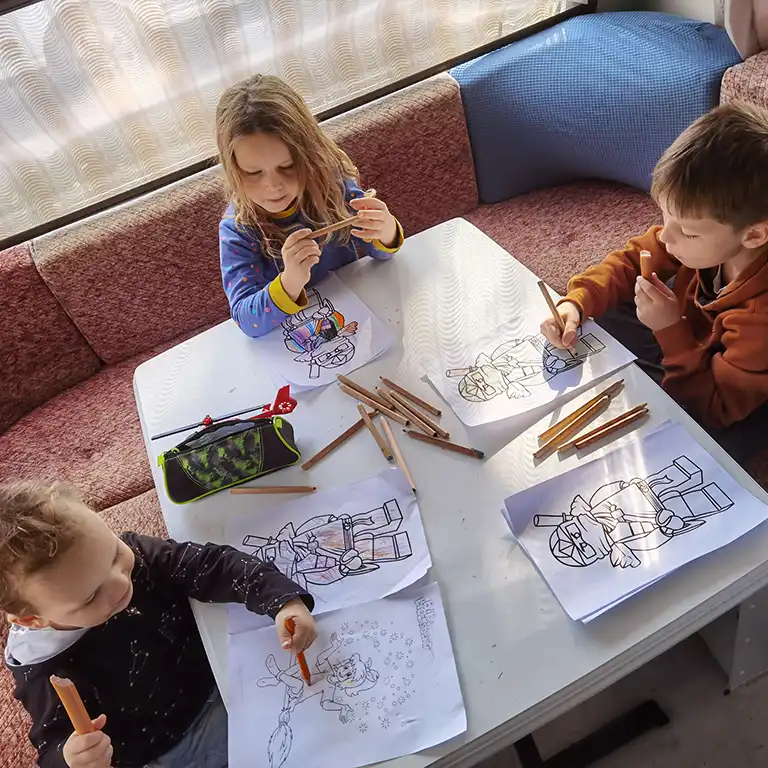 Three children coloring pictures with colored pencils at a table inside a caravan during a holiday at Südsee-Camp