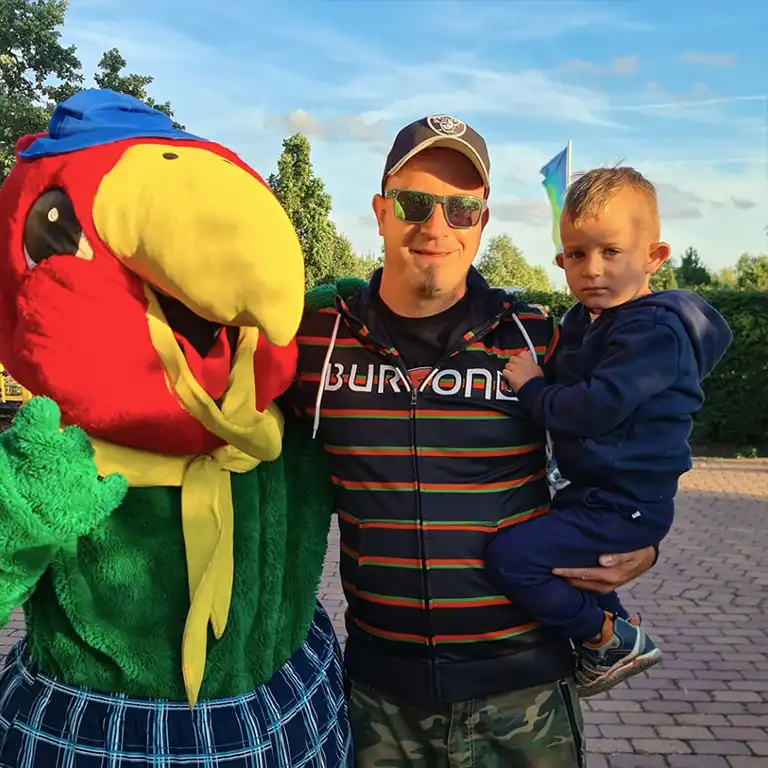 Father with young child posing next to Südsee-Camp mascot Campino, a colorful parrot in costume