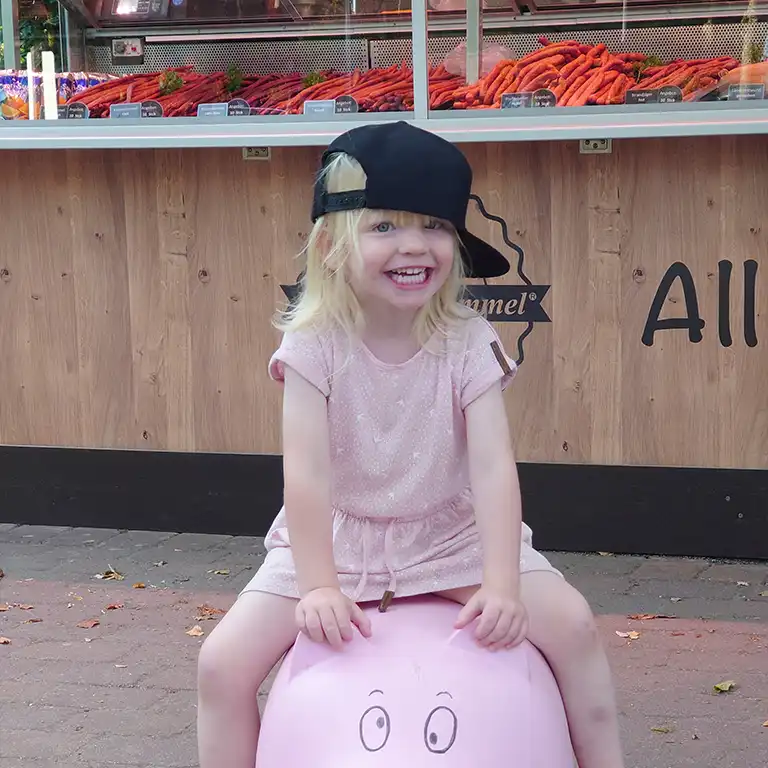 Smiling blonde child wearing a black cap sits on a pink pig toy in front of the Wurstkult sausage stand at Südsee-Camp
