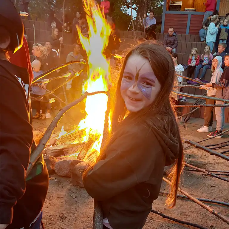 A girl with colorful face paint smiles at the camera while holding a stick with dough over the campfire – ready to make delicious bread on a stick. In the background, families sit together and enjoy the evening. A real highlight for kids at Südsee-Camp.
