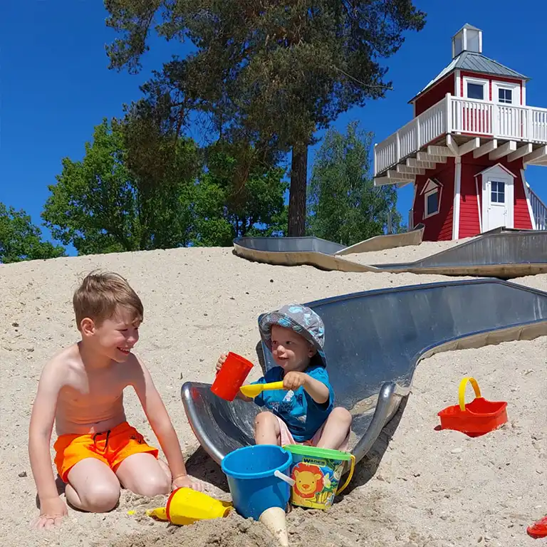 Two children happily play in the sand at the lighthouse playground at Südsee-Camp. On a relaxing family day, they enjoy the sunshine, slide down the slide, and build with colorful sand toys – perfect for little explorers.
