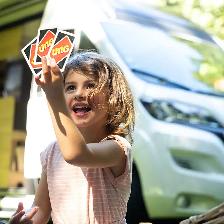 A cheerful child happily plays UNO in front of a camper van. The image captures carefree family moments and relaxed camping surrounded by nature.