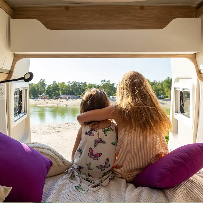 Two children sit close together in the open camper van, looking out over the idyllic lake at Südsee-Camp. The peaceful scenery and view of the sandy shore capture the essence of a perfect family holiday in nature.