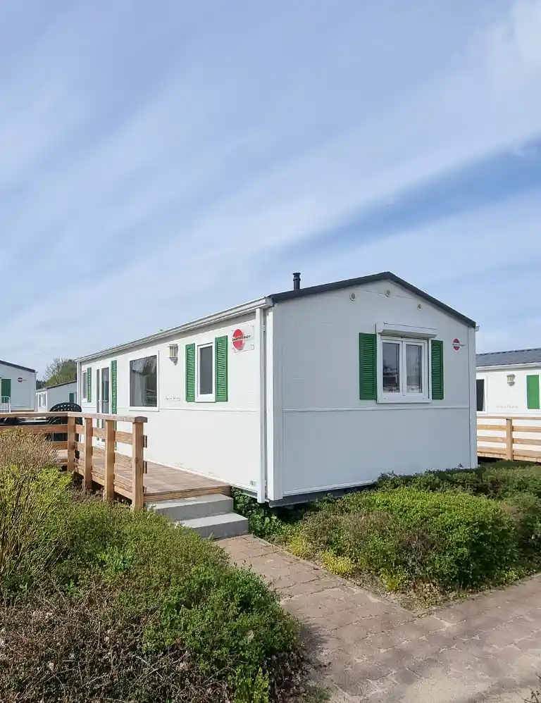 White Type 1 chalet at Südsee-Camp with green shutters, a wooden terrace with ramp and railing, surrounded by low shrubs and a paved path. The sky is bright with light, wispy clouds.