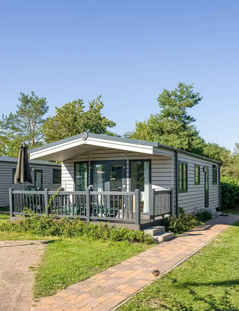 Modern mobile home Type 2+ at Südsee-Camp with covered terrace and garden furniture, surrounded by well-kept lawn and trees under a clear blue sky.