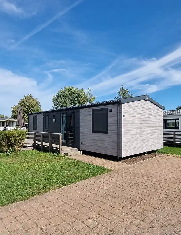 Modern Type 3 chalet at Südsee-Camp with light wooden exterior and dark window frames, a small wooden terrace with ramp, green lawn, and paved path. The sky is blue with light wispy clouds.
