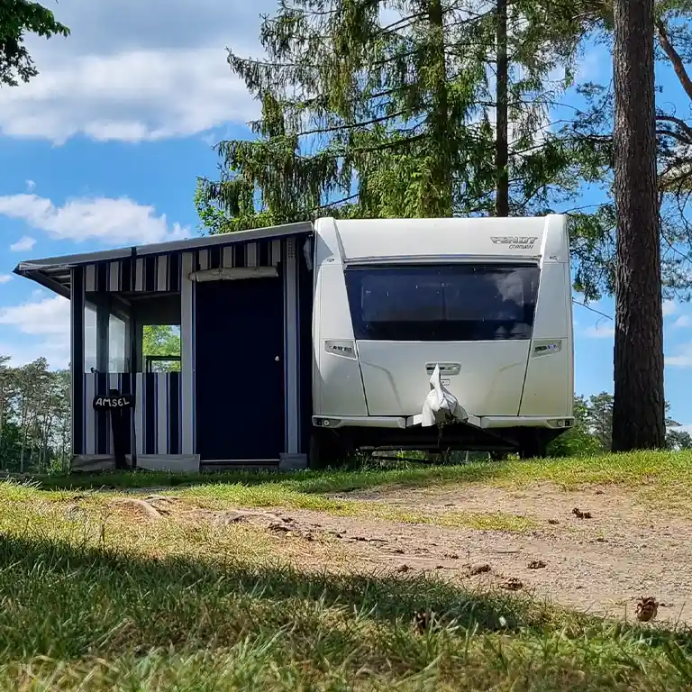 go to rental caravans Rental caravan at Südsee-Camp with a fixed striped awning on a natural pitch beneath tall trees, under a blue sky with scattered clouds.