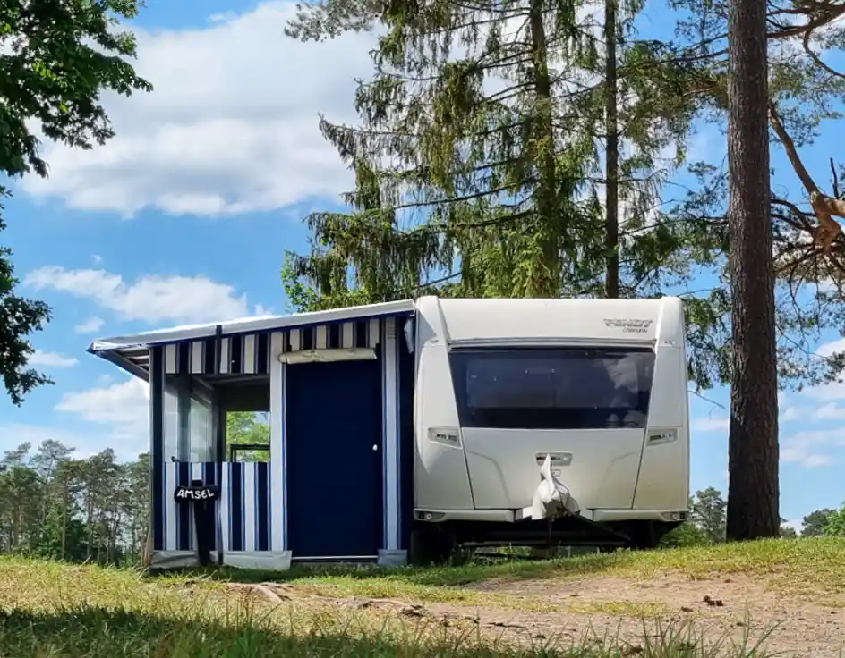 Caravan type 3 at Südsee-Camp White caravan with a fixed blue-and-white striped awning labeled “Amsel,” located on a green pitch at Südsee-Camp. Surrounded by pine trees under a blue sky with light clouds.