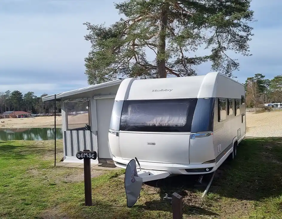 Wohnwagen Typ 4 im Südsee-Camp Weißer Mietwohnwagen mit festem Vorzelt im Südsee-Camp, direkt am Wasser gelegen. Im Vordergrund ein Holzschild mit der Aufschrift „GEMSE“ und eine Satellitenschüssel. Im Hintergrund sieht man einen Sandstrand und Bäume.