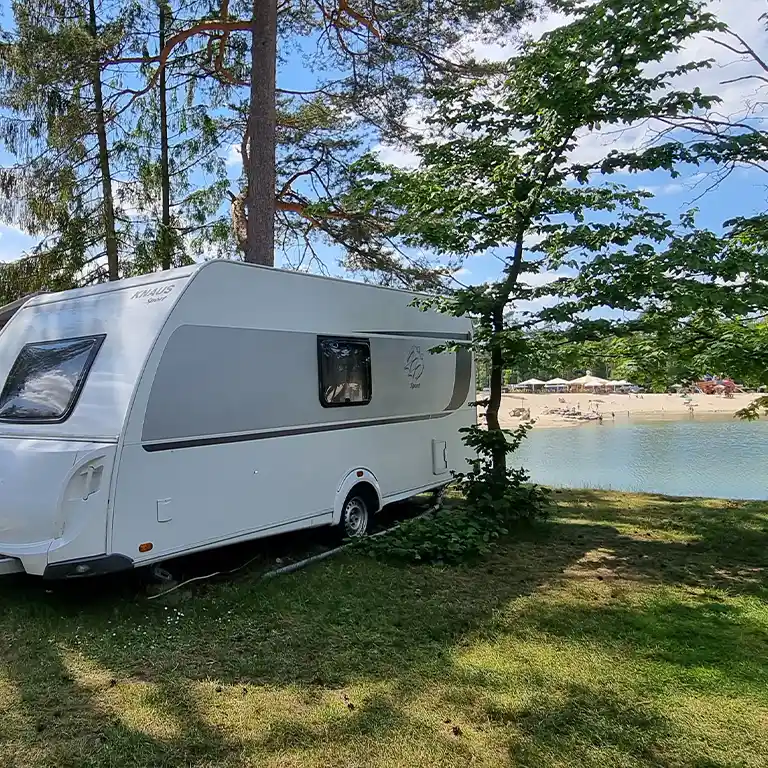 Weißer Wohnwagen vom Typ Knaus Sport auf einem schattigen Stellplatz direkt am Ufer des Badesees im Südsee-Camp. Im Hintergrund sieht man einen Sandstrand mit Sonnenschirmen und Badegästen unter blauem Himmel mit wenigen Wolken.