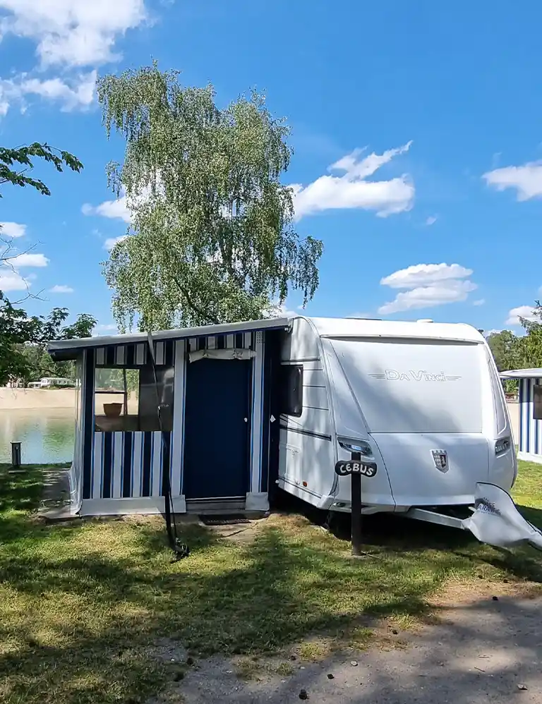 Weißer Mietwohnwagen mit blau-weiß gestreiftem festem Vorzelt im Südsee-Camp. Im Hintergrund ist ein See mit Sandstrand zu sehen. Vor dem Wohnwagen steht ein Schild mit der Aufschrift „Cebus“. Ein großer Baum spendet teilweise Schatten.