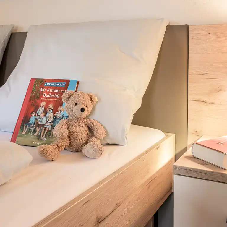 Parents' bed in a Swedish-style holiday house at Südsee-Camp – a light brown teddy bear and the children's book "The Children of Noisy Village" by Astrid Lindgren lie on the bed, creating a cozy, family-friendly atmosphere.