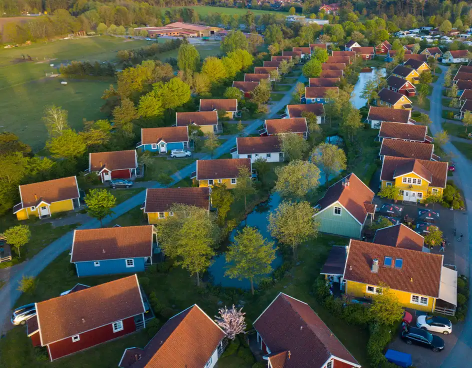 Aerial view of the colorful Scandinavian-style holiday homes at Südsee-Camp, surrounded by ponds, trees, and paths – a picturesque holiday village near open meadows at sunset.